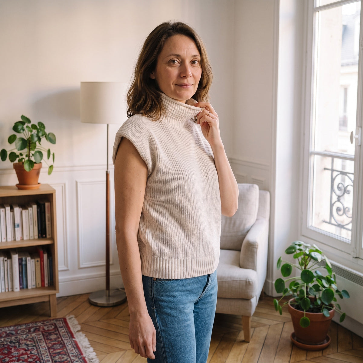 Le même pull en blanc cassé, portée de trois quarts près de la fenêtre, une main qui remonte le col montant