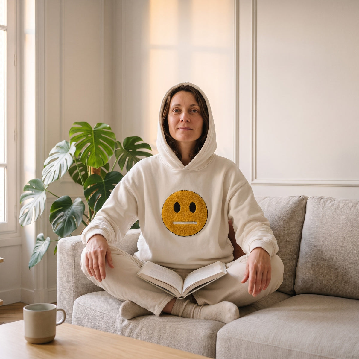 Femme assise en tailleur sur son canape, un livre ouvert devant elle, en sweat blanc creme capuche relevee, le smiley jaune au centre de la poitrine