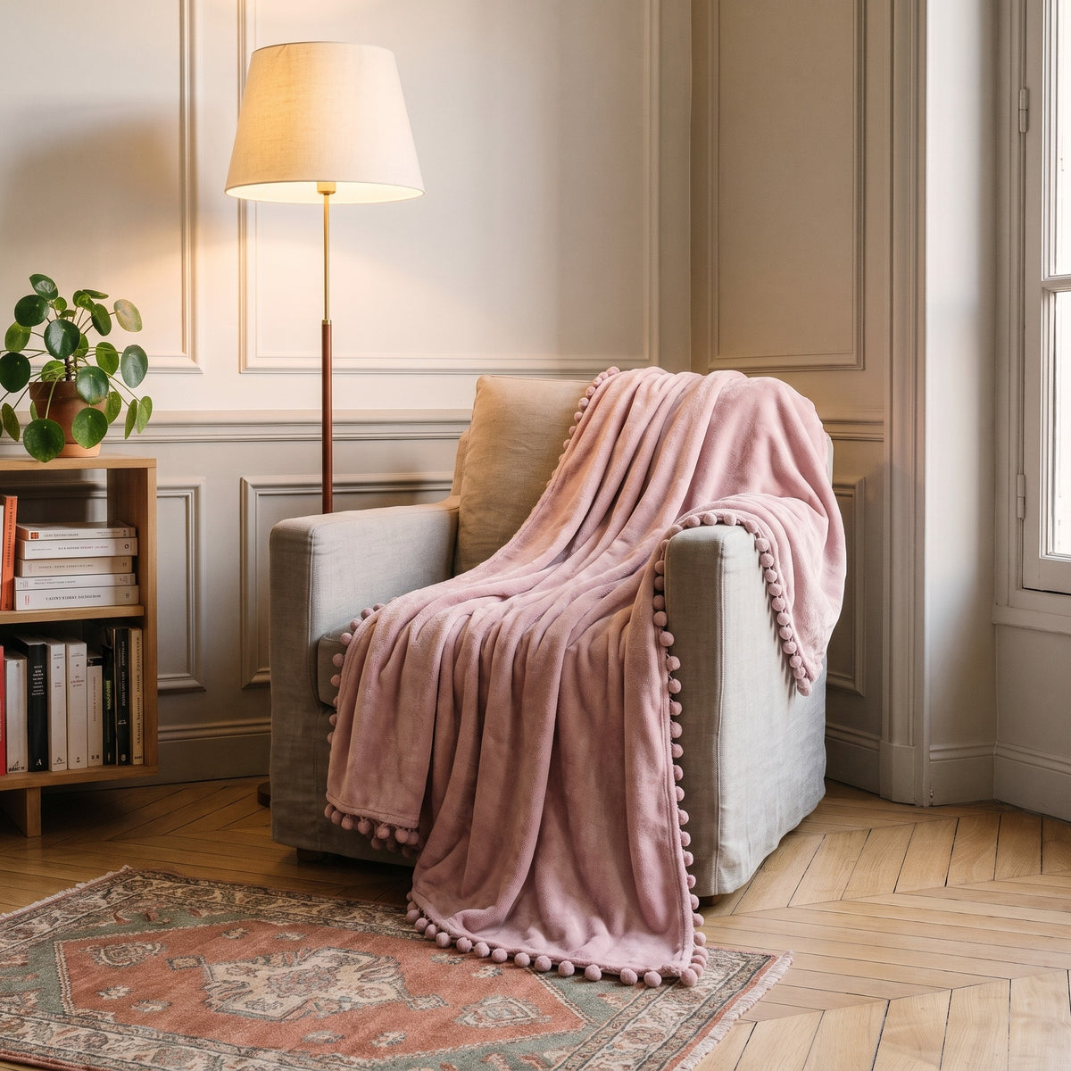 Le plaid rose poudré posé sur un fauteuil de lecture dans un salon haussmannien, un angle retombe sur l’accoudoir et un pan touche le tapis, le lampadaire est allumé en fin de journée