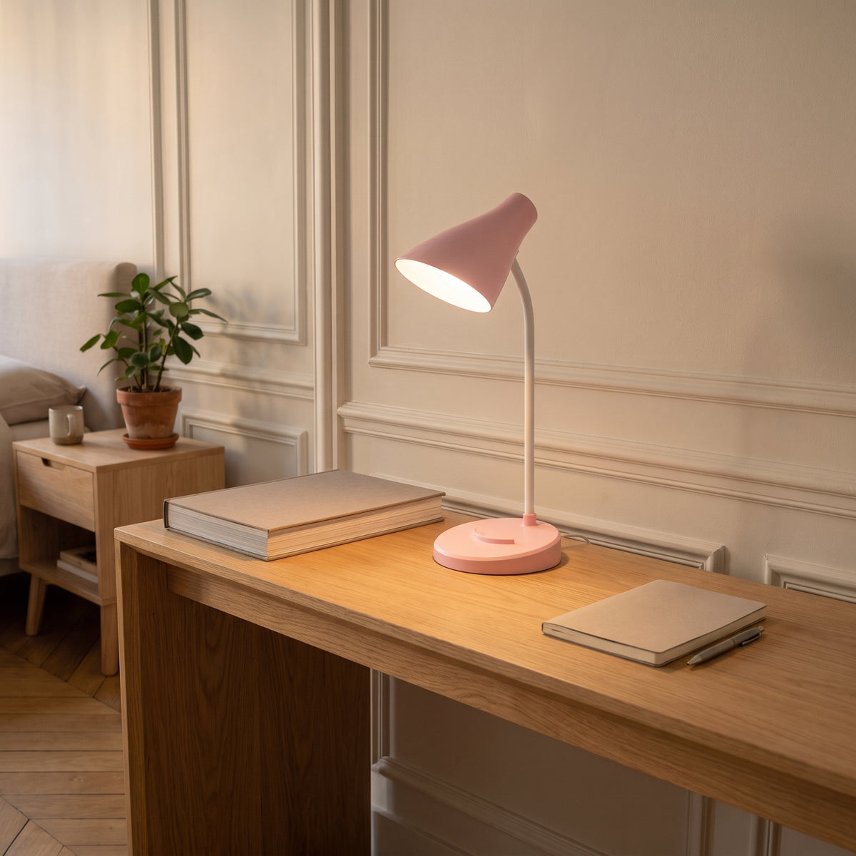 La lampe rose allumée au bord d'une console en chêne, un gros livre relié à sa gauche et un carnet à sa droite, la lumière étalée sur le bois.