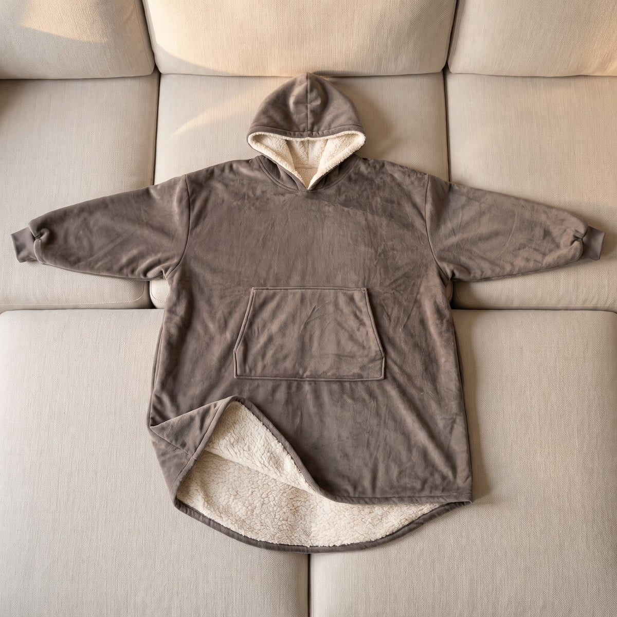 Le plaid à capuche gris taupe étalé à plat sur un canapé, vu de dessus : capuche, grande poche du devant, poignets resserrés, et le coin du bord bas retourné sur la doublure sherpa
