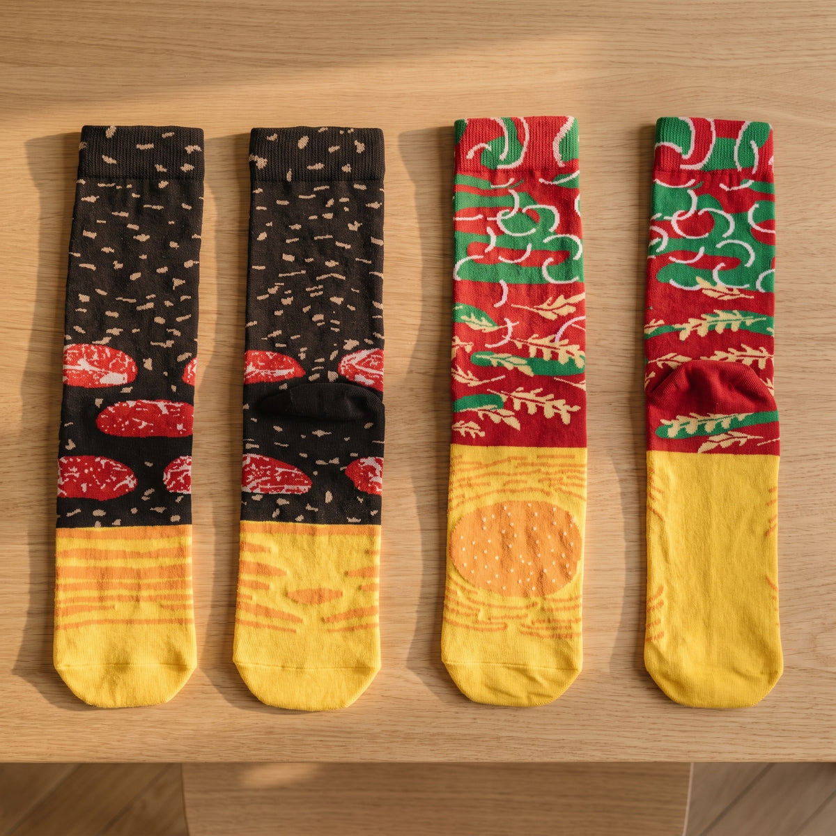 Les quatre chaussettes du coffret posées à plat côte à côte sur une table en chêne clair : deux au fond brun semé de graines et de tranches rouges, deux rouges et vertes à feuilles beiges, toutes avec le pied jaune