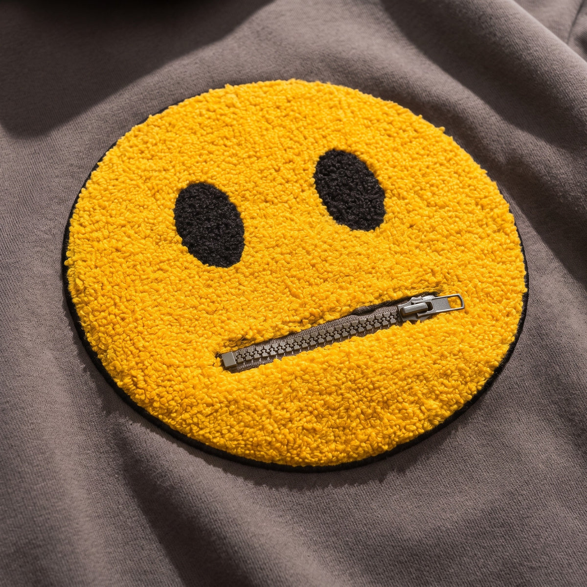 Le smiley vu de tres pres en lumiere rasante : le fil jaune boucle en relief, les deux yeux noirs dans la meme matiere, et la glissiere metallique qui lui fait la bouche