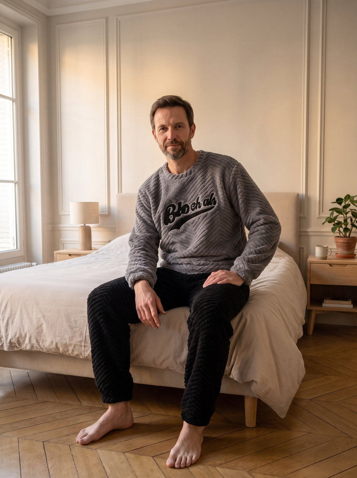 Homme assis au bord du lit en pyjama polaire gris et noir, le mot Blochah brode en noir sur la poitrine.