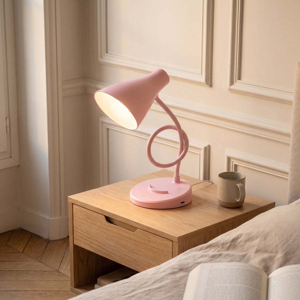 La lampe rose sur une table de chevet, le bras enroulé en boucle : l'abat-jour est descendu de plusieurs centimètres et se penche vers la gauche.