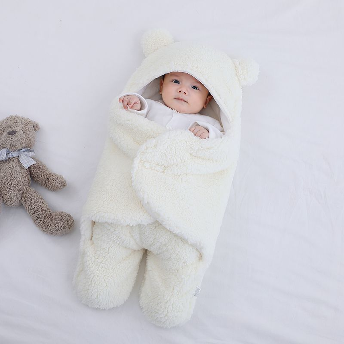 Bebe dans la gigoteuse creme en polaire bouclee, capuche relevee, un ourson en peluche gris pose contre lui