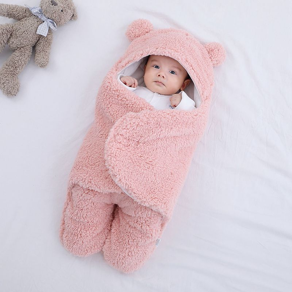 Bebe dans la gigoteuse rose clair en polaire bouclee, capuche relevee avec ses deux oreilles rondes, un ourson en peluche a cote