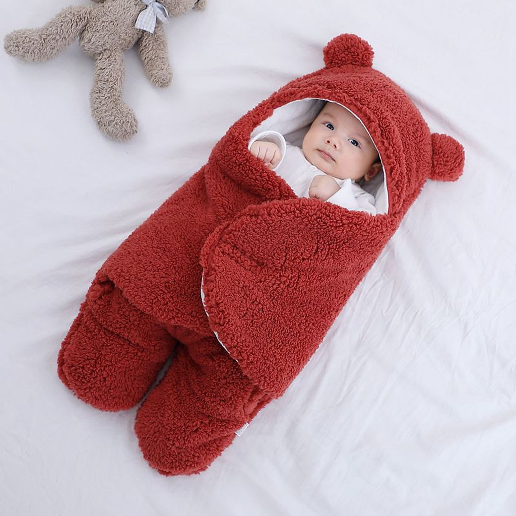 Bebe dans la gigoteuse rouge brique, capuche relevee avec ses deux oreilles rondes, un ourson en peluche pose a cote sur le drap