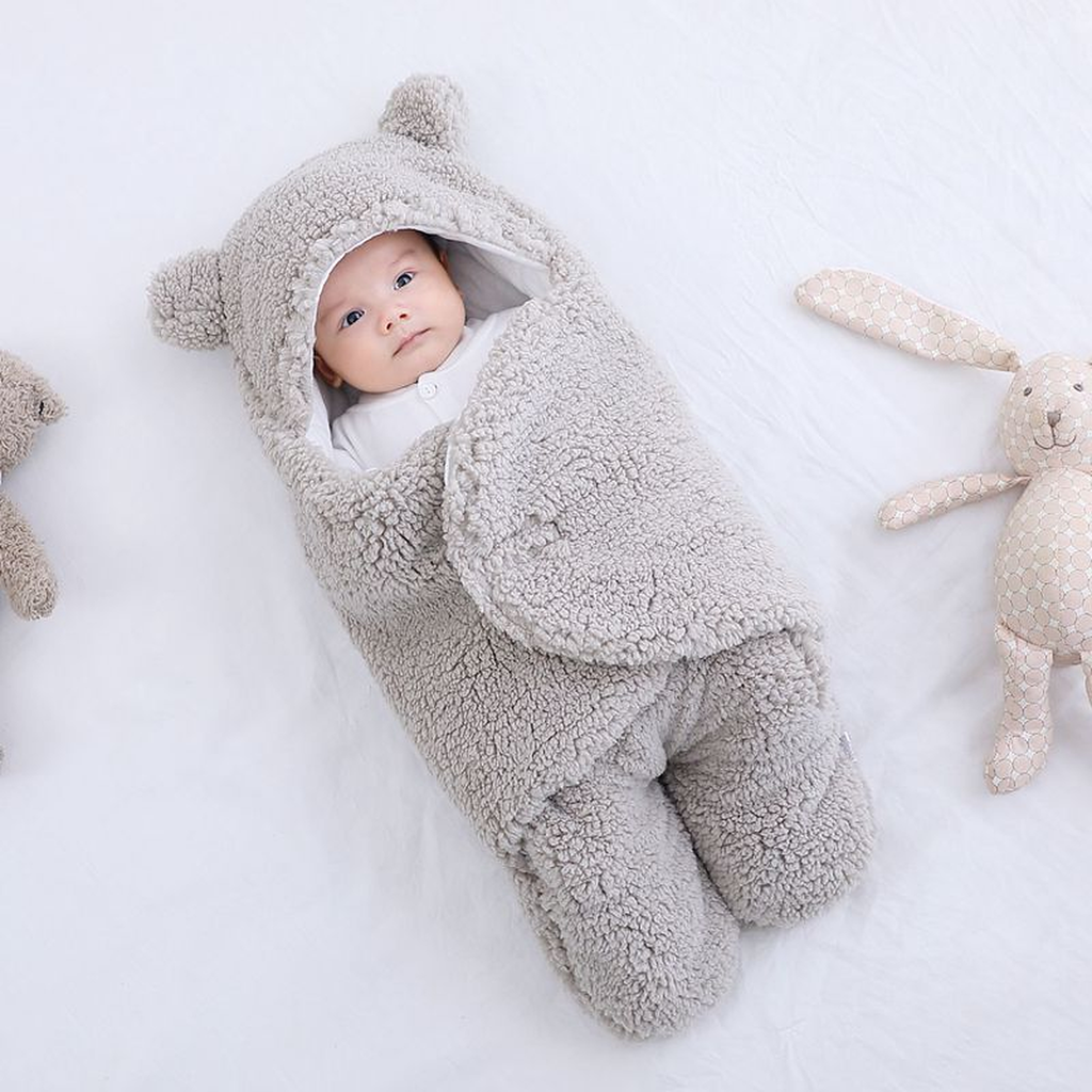 Bebe dans la gigoteuse gris clair en polaire bouclee, capuche relevee, un lapin en peluche couche a cote de lui