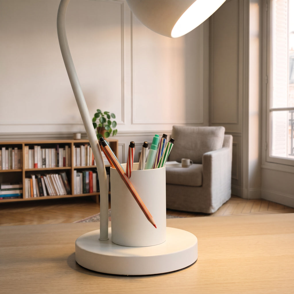 Plan rapproché du pied de la lampe blanc crème : le godet garni de stylos, un crayon couché dans l'encoche de sa paroi, et le socle rond posé sur le bois