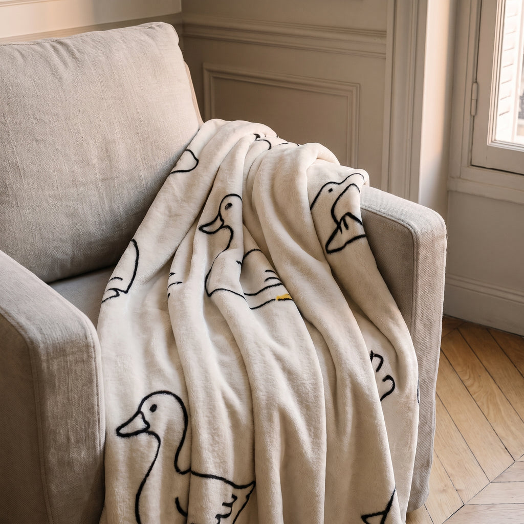 Plaid polaire à motifs, modèle Canards écru, posé sur un fauteuil de lecture