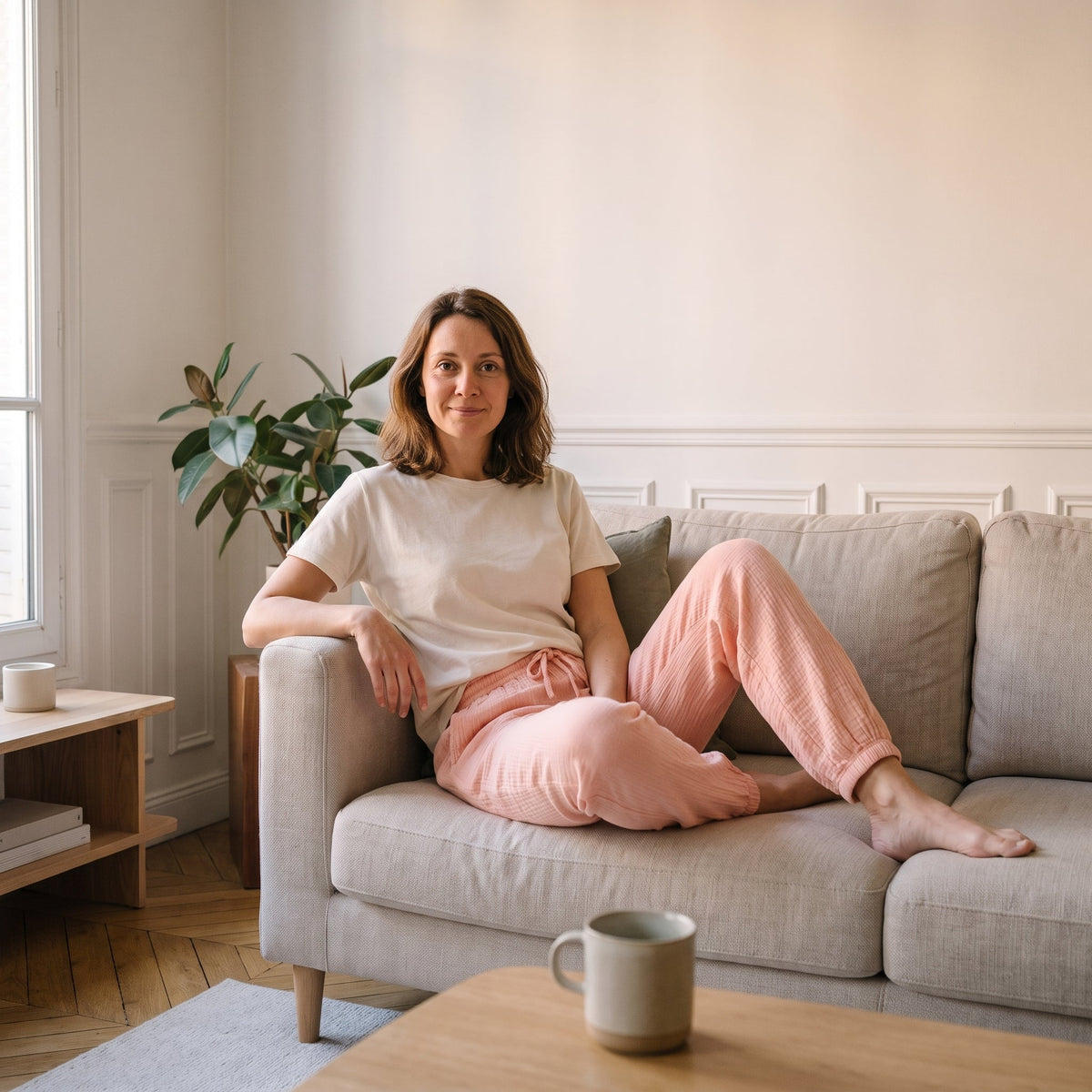 Femme assise sur son canape en pantalon de pyjama saumon en gaze de coton et tee-shirt ecru