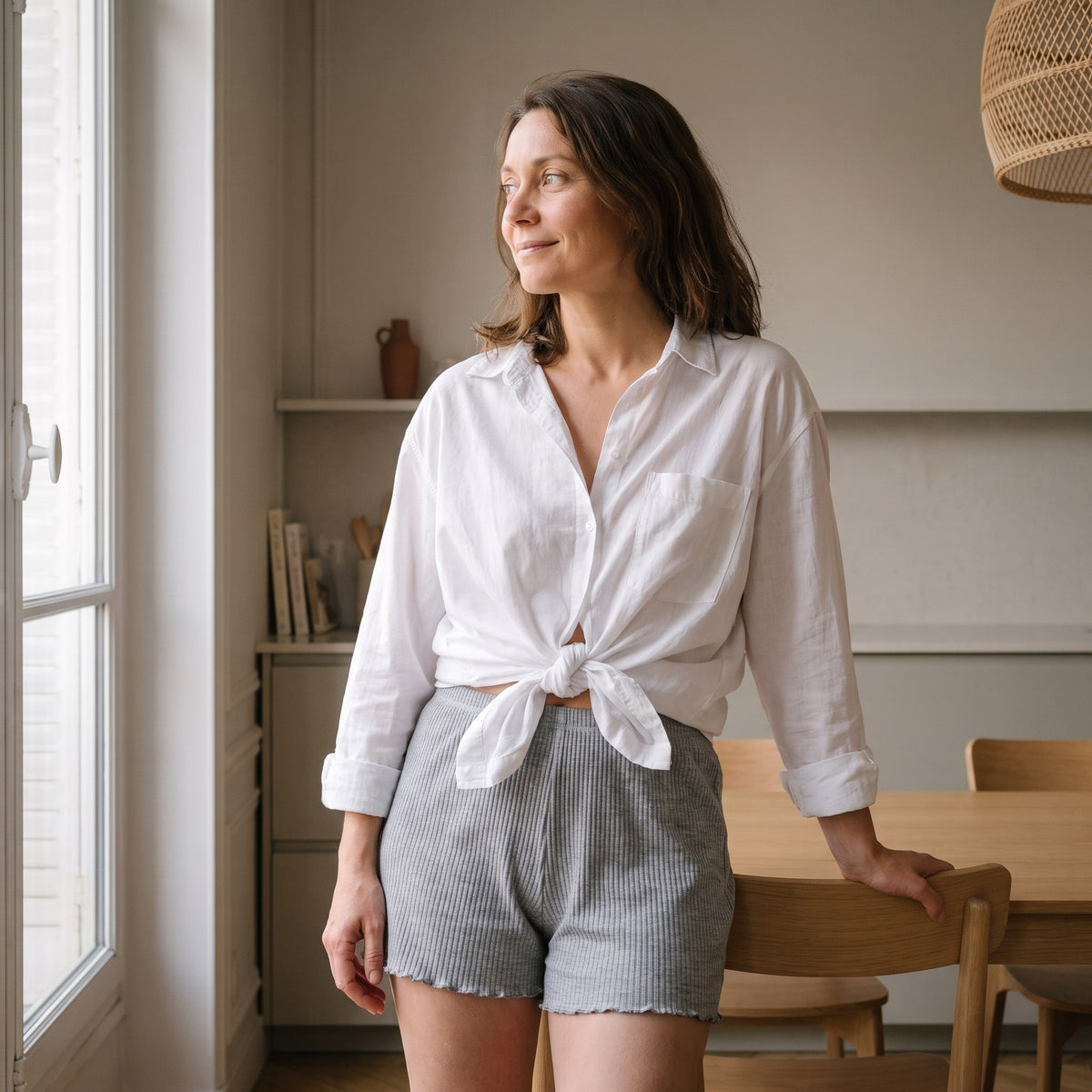 Femme en chemise blanche portant le short boxer gris, debout près de la fenêtre du coin repas