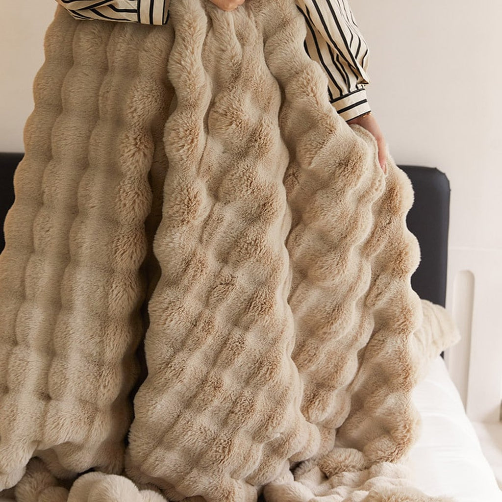 Plaid beige imitation fourrure tenu devant un lit, montrant sa largeur, sa retombée et ses rangées en relief