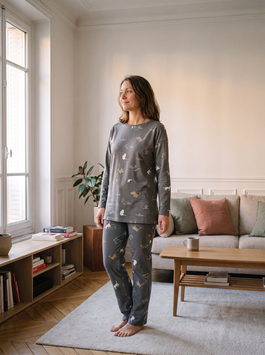 Femme portant l ensemble pyjama gris-vert seme de petits chats, debout dans un salon, haut et pantalon assortis.