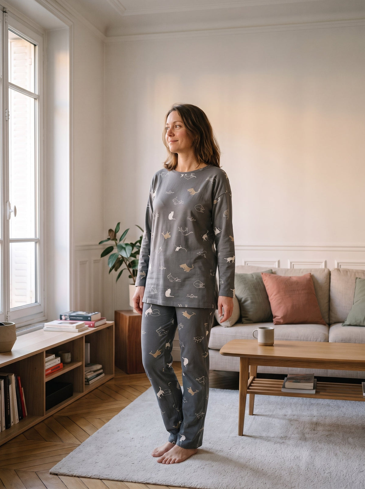 Femme portant l ensemble pyjama gris-vert seme de petits chats, debout dans un salon, haut et pantalon assortis.