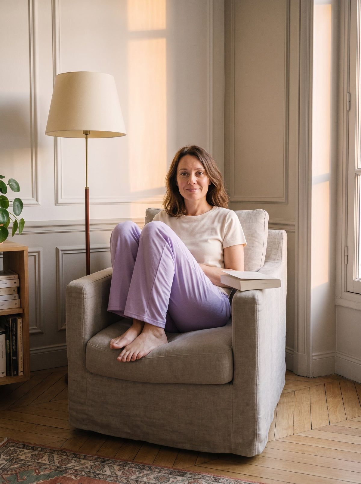 Bas de pyjama lilas porte dans le fauteuil, jambes repliees de cote, un livre a la main.