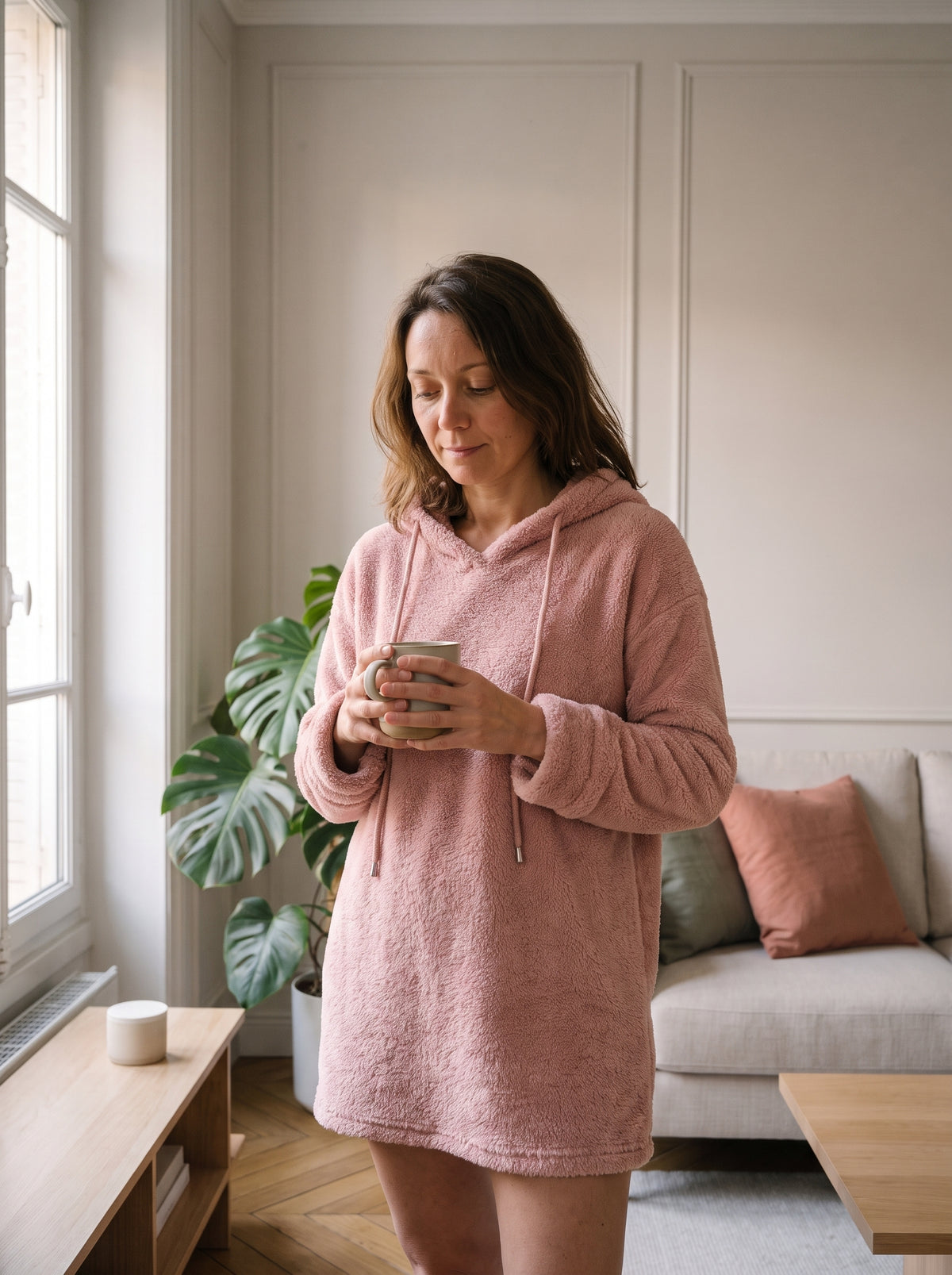 Femme du casting portant le pull long à capuche peluche rose près de la fenêtre du salon