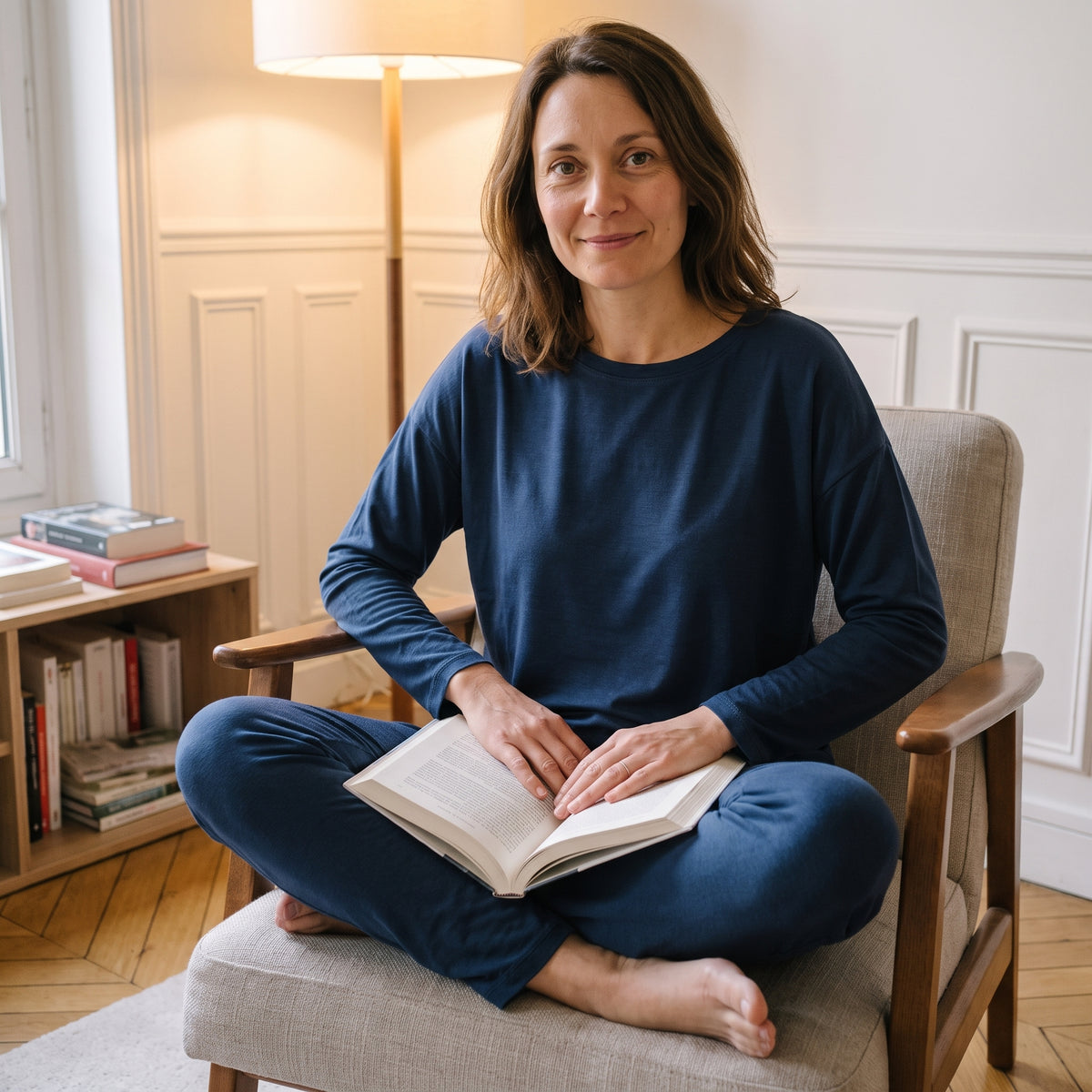 Femme assise en tailleur dans un fauteuil de lecture, en ensemble pyjama modal bleu marine, un livre ouvert.