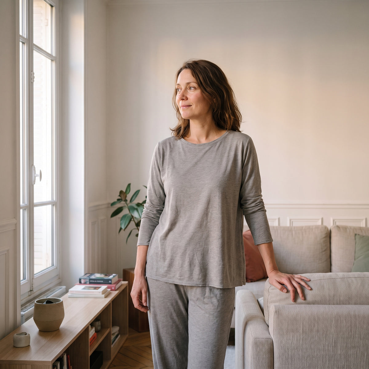 Femme debout près de la fenêtre du salon en ensemble pyjama modal gris taupe, haut ample à ourlet droit et bas assorti.