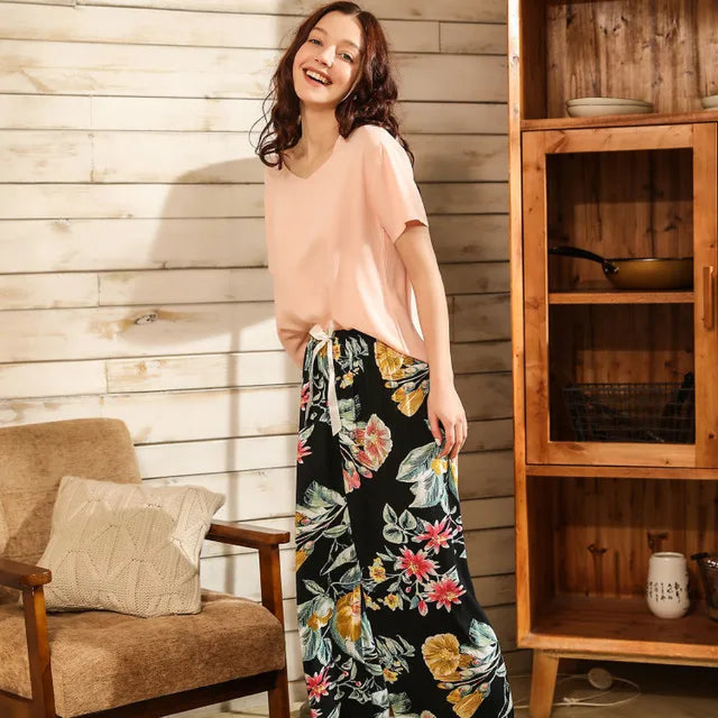 Pyjama femme, haut rose pâle et pantalon large fleurs noir, debout devant une cloison en bois clair, le buste tourné.