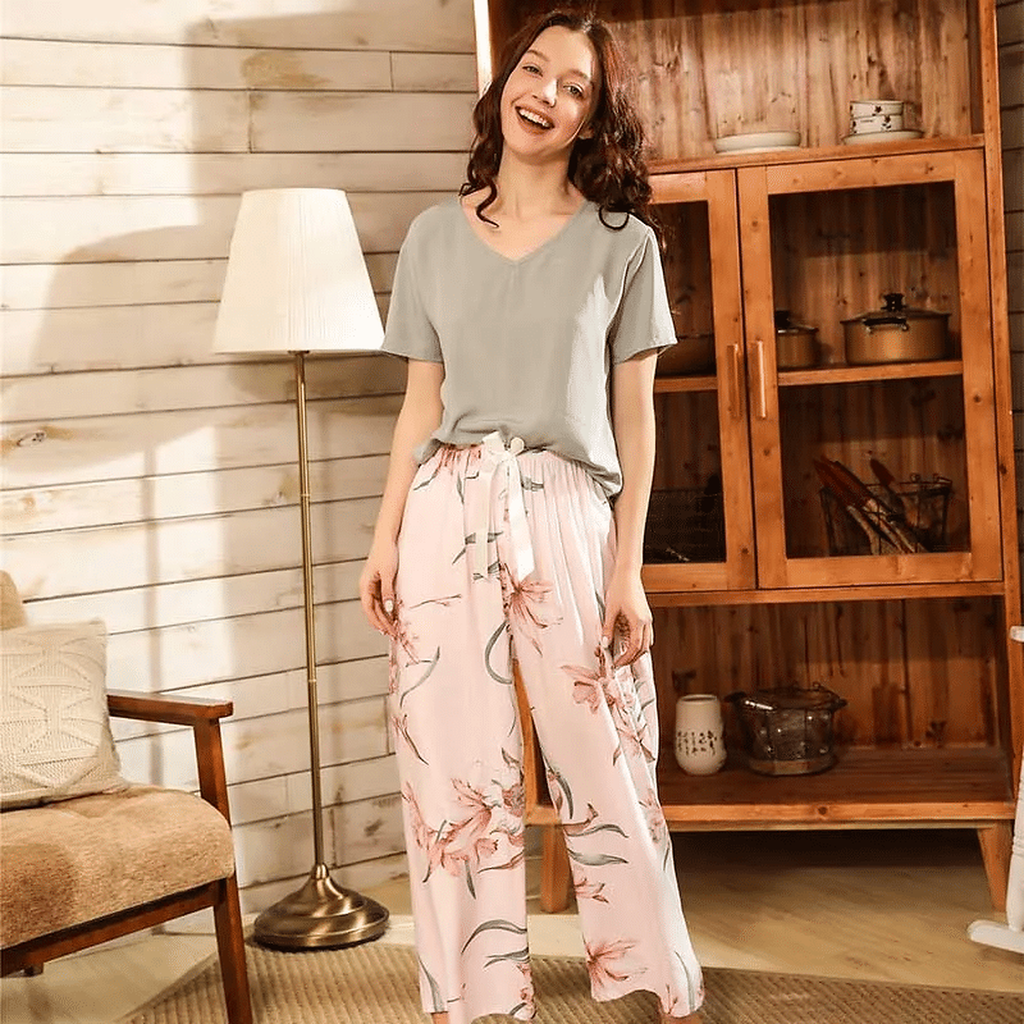 Pyjama femme, haut sauge et pantalon large magnolias rose, debout devant un vaisselier en bois, la main sur la hanche.