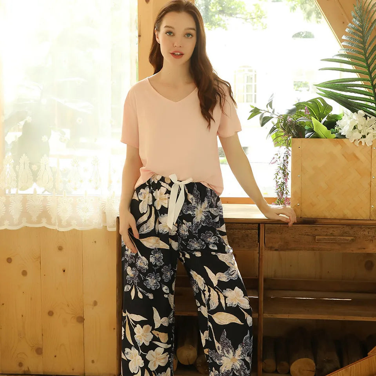 Pyjama femme, haut beige rosé et pantalon large feuillage doré marine, debout devant une fenêtre, à contre-jour.