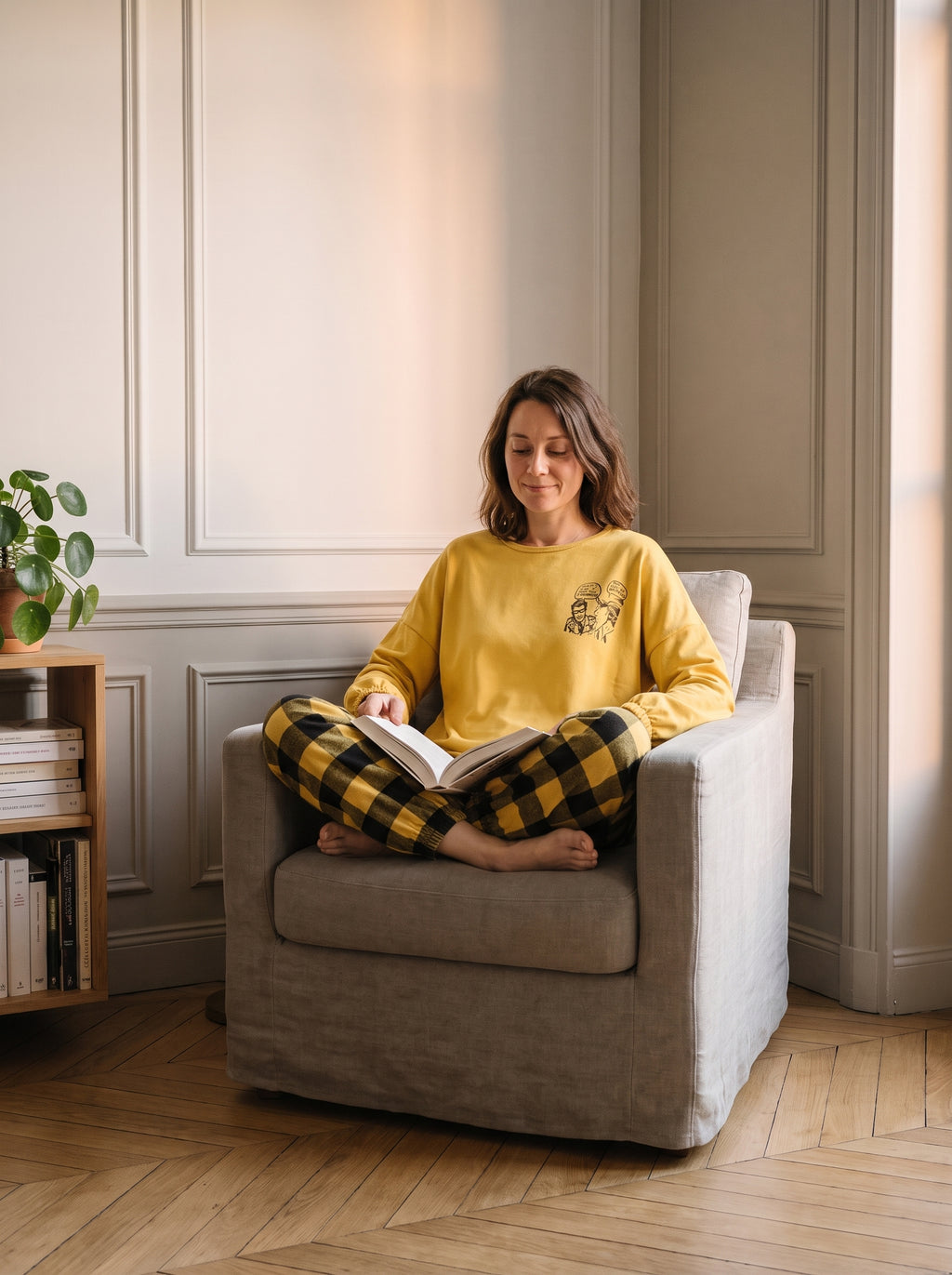 Femme assise en tailleur dans un fauteuil, un livre ouvert, en pyjama jaune à carreaux