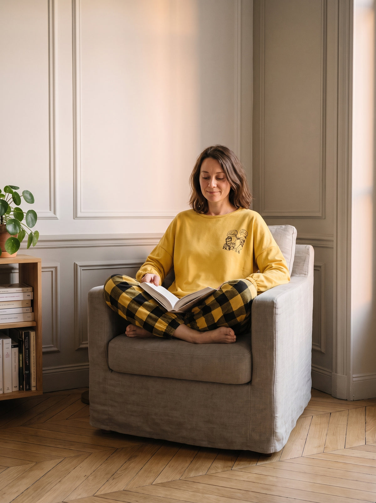 Femme assise en tailleur dans un fauteuil, un livre ouvert, en pyjama jaune à carreaux