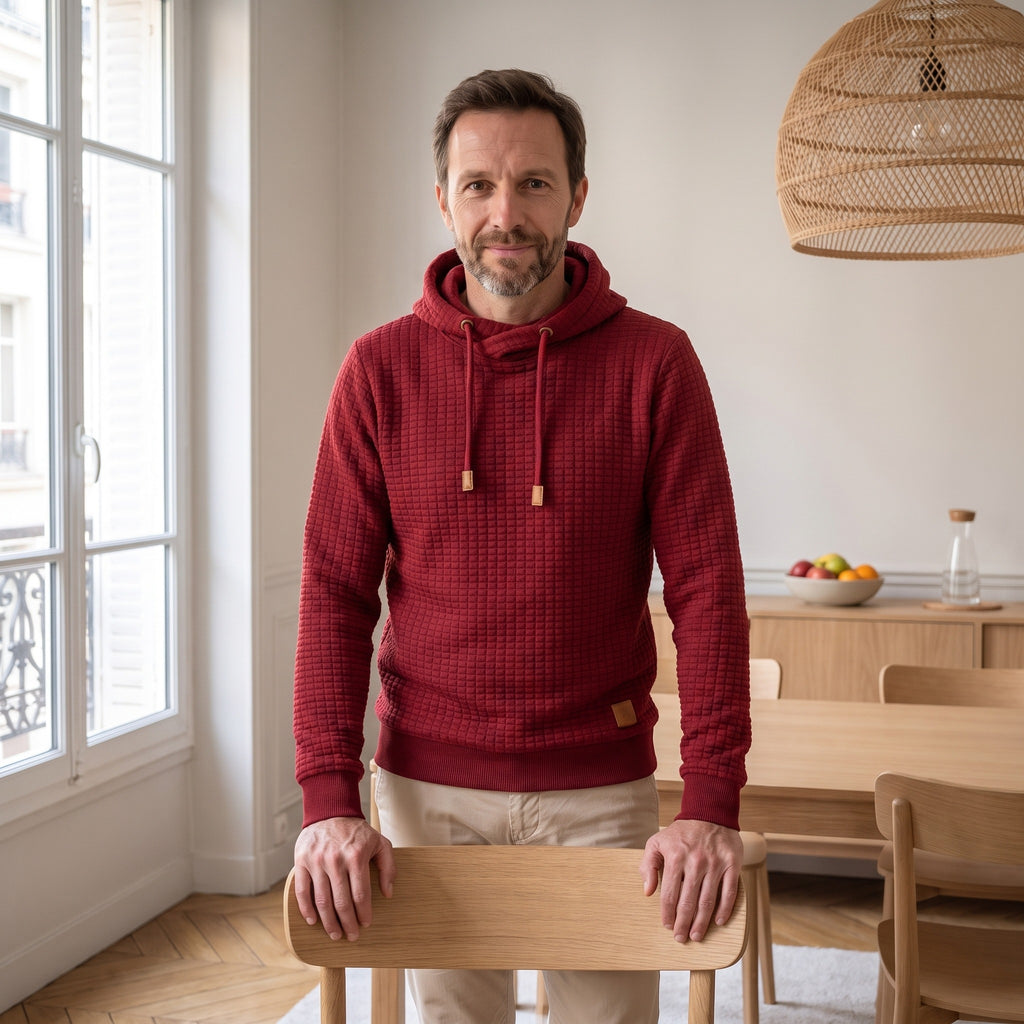 Sweat à capuche gaufré rouge porté de face, les deux mains posées sur le dossier d'une chaise du coin repas.