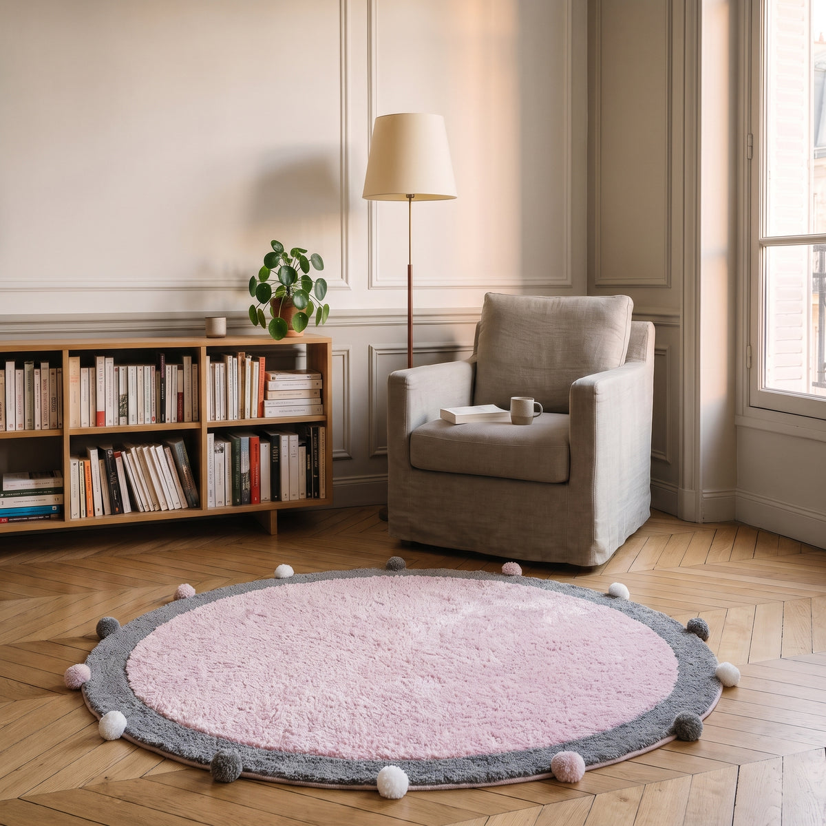 Tapis rond à pompons rose au sol d'un coin lecture, à côté d'un fauteuil et d'une étagère qui donnent l'échelle