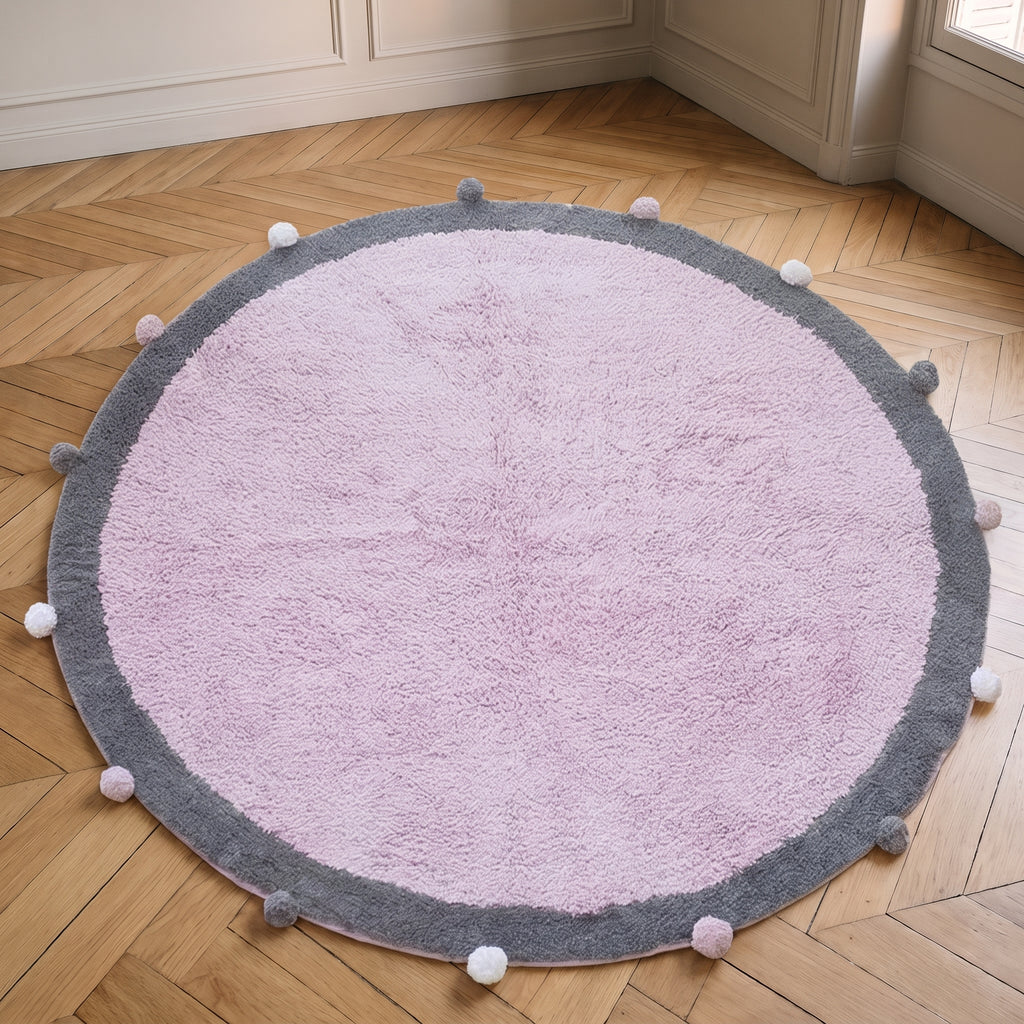Tapis rond à pompons rose vu de dessus, posé à plat sur le sol d'une pièce claire, le cercle entier dans le cadre
