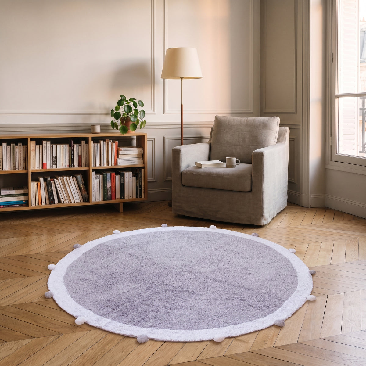 Tapis rond à pompons gris perle posé au sol d'un coin lecture, près d'un fauteuil et d'un lampadaire