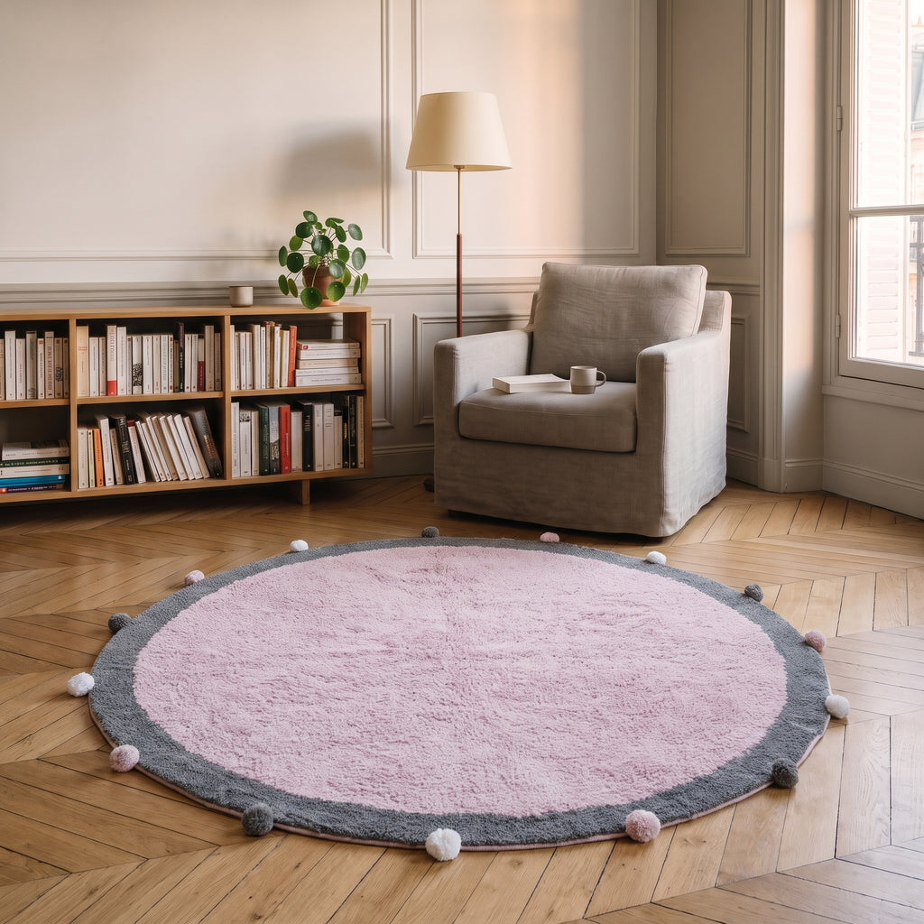 Tapis rond à pompons rose posé au sol d'un coin lecture, au pied d'un fauteuil, vue en perspective