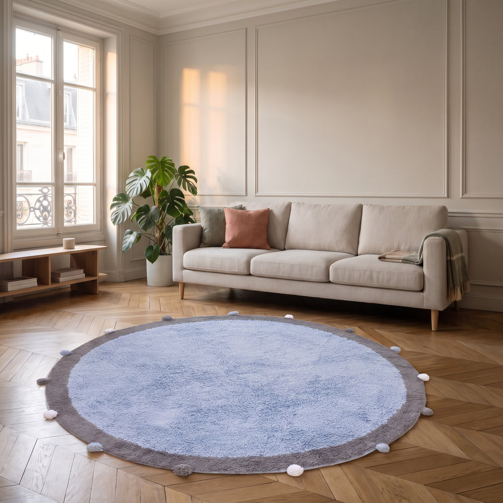 Tapis rond à pompons bleu ciel posé au sol d'un salon lumineux, devant un canapé clair