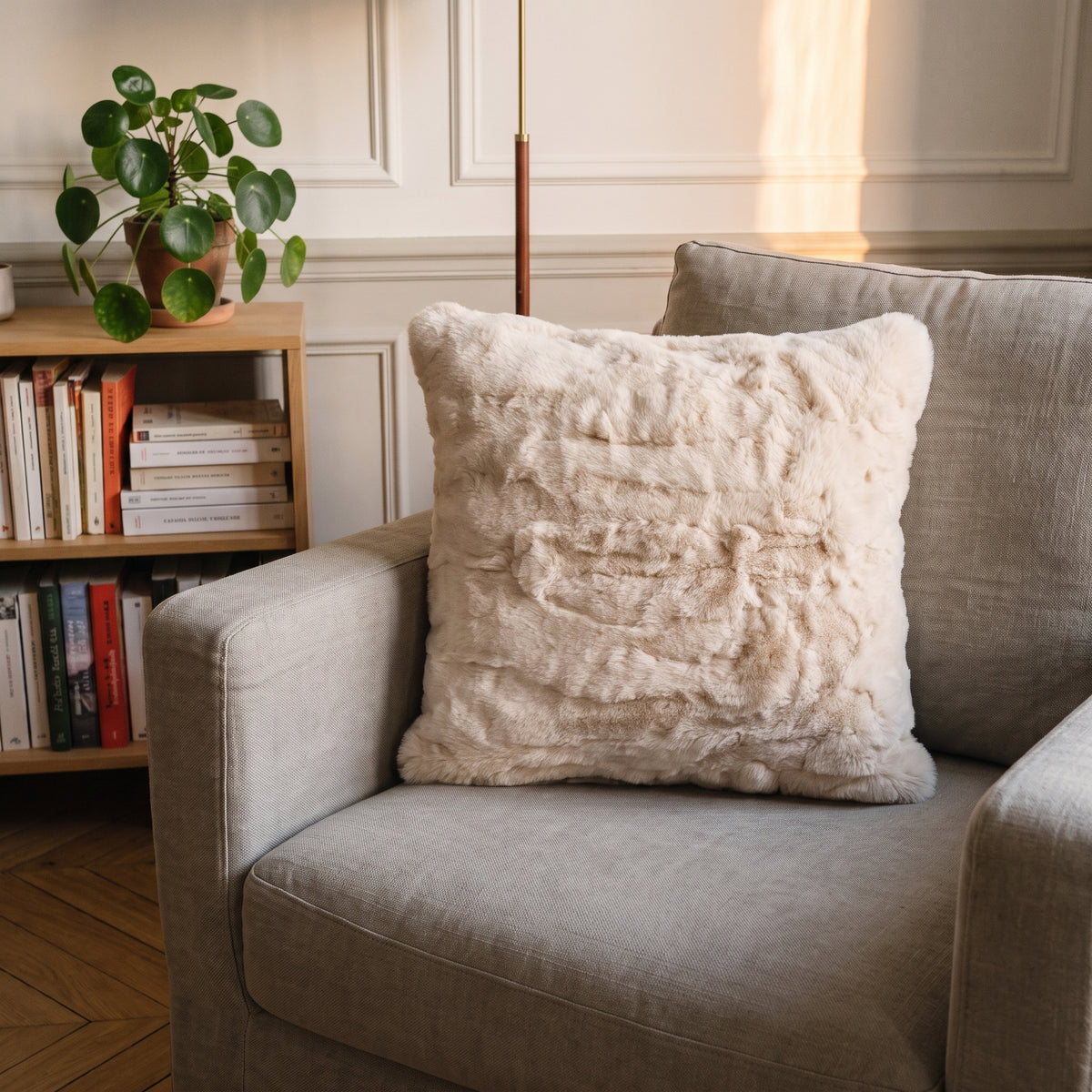 Coussin extra doux en fausse fourrure beige calé dans un fauteuil, devant une bibliothèque
