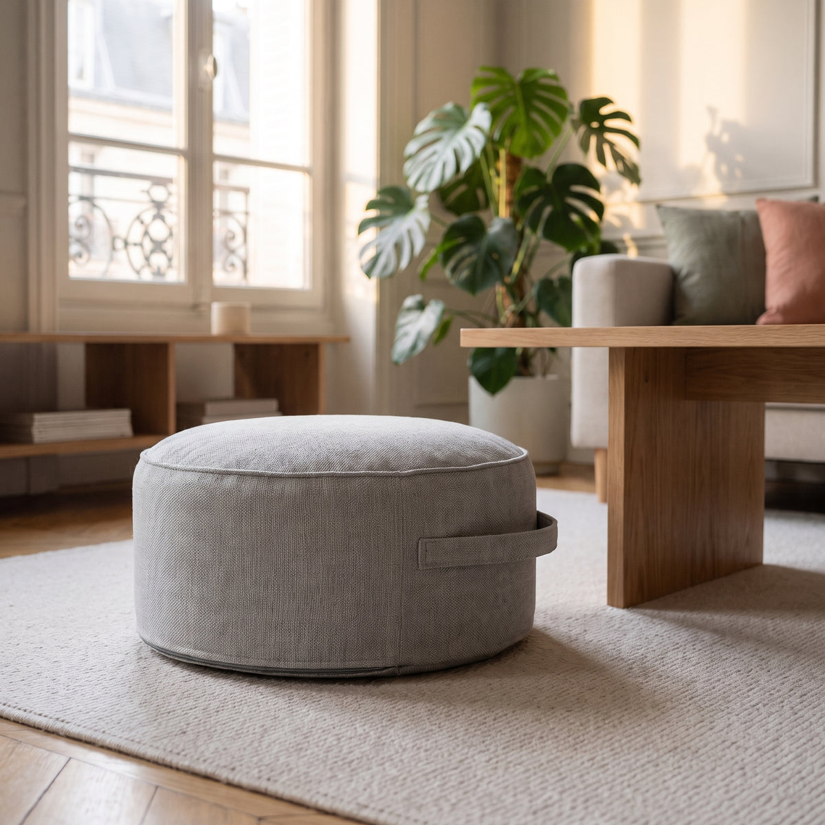 Pouf de sol rond gris perle posé sur le tapis du salon, poignée visible