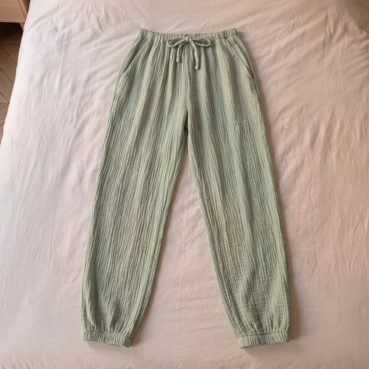 Pantalon de pyjama vert sauge en gaze de coton pose a plat sur la literie, vu du dessus